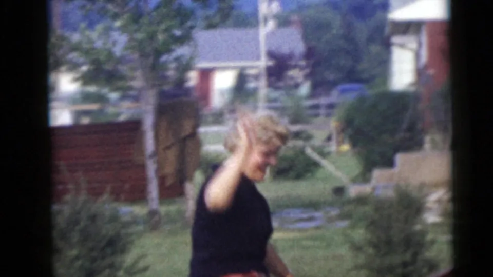 The woman smiles and raises her hand in a wave while walking outdoors, with houses, grass, trees, and shrubs in the background.