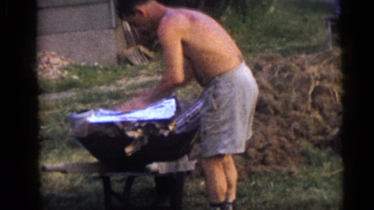 Shirtless man bends over a wheelbarrow in a grassy backyard.