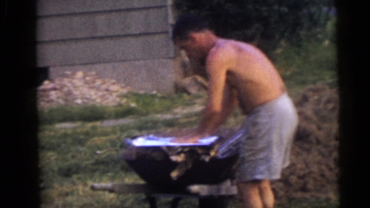 A shirtless man in light shorts works over a wheelbarrow in a Camden backyard, with grass and a dirt pile nearby.