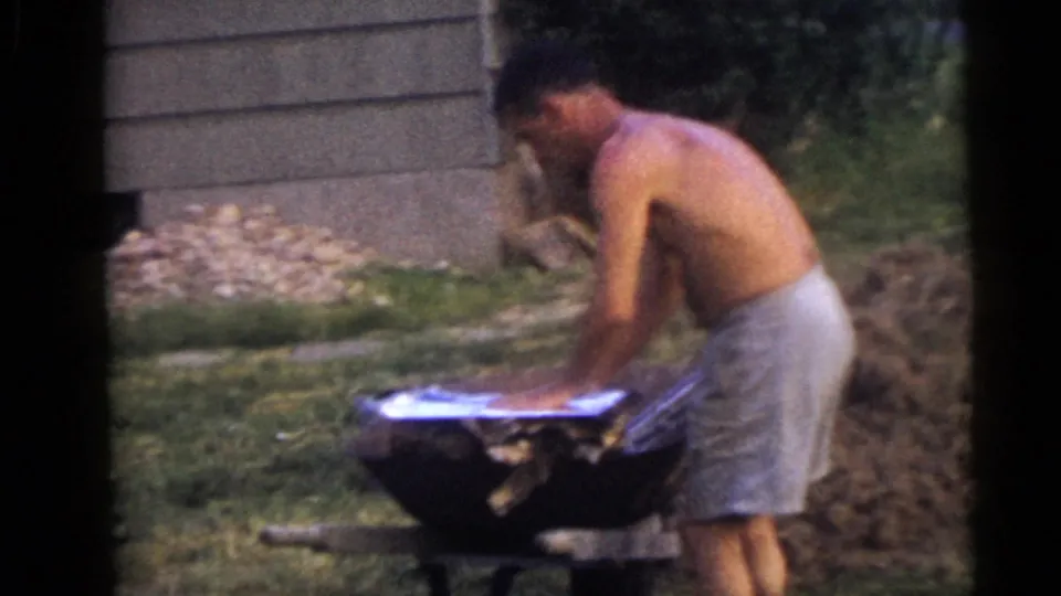 A shirtless man in light shorts works over a wheelbarrow in a Camden backyard, with grass and a dirt pile nearby.