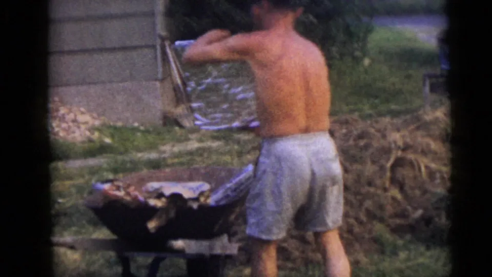 The man stands with his back to the camera while lifting or adjusting sheet material near the wheelbarrow in the yard.
