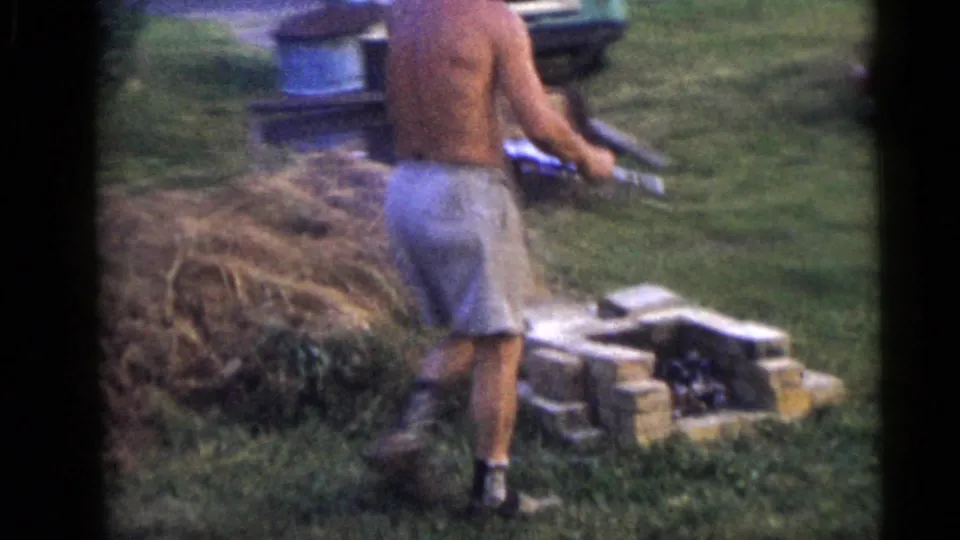 The man walks away from the wheelbarrow carrying sheet material near a dirt pile and small brick or block fire area.