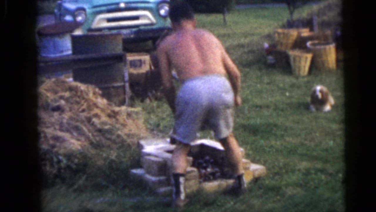 Shirtless man bends over a small brick structure in a grassy yard near a vintage truck.