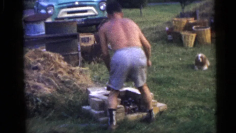 In 1960 Camden home-movie footage, a shirtless man works at a small brick structure in a yard with hay and a green truck nearby.