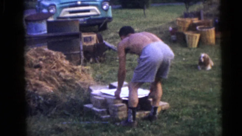 A shirtless man in shorts works over a low brick structure in the yard, framed by hay, a vintage truck front, and a small dog.
