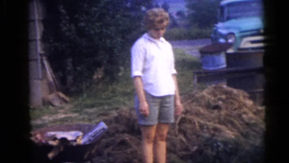 A woman in a white shirt and shorts stands beside the yard work area, with hay, a wheelbarrow, and a green truck visible nearby.
