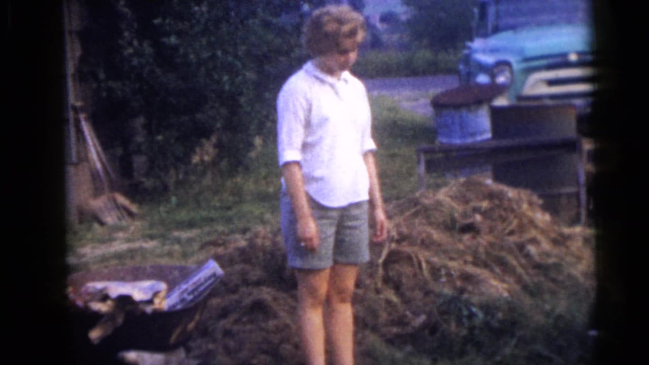 Woman in a white shirt and shorts stands in a grassy yard near branches and a teal parked car.