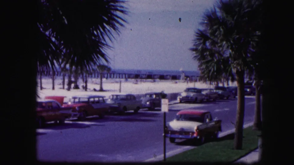 1960 Miami beach view with palm trees, parked classic cars, bright sand, ocean, and a distant pier seen through dark foreground framing.