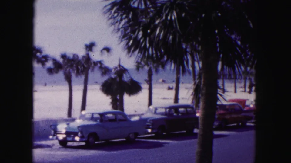 1960 Miami beach road with classic cars in the foreground, palm trees, scattered beachgoers, sand, and ocean beyond.