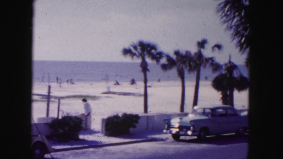 1960 Miami beach scene with a man near a parked vehicle, palm trees, classic cars, beach sand, and ocean in the background.