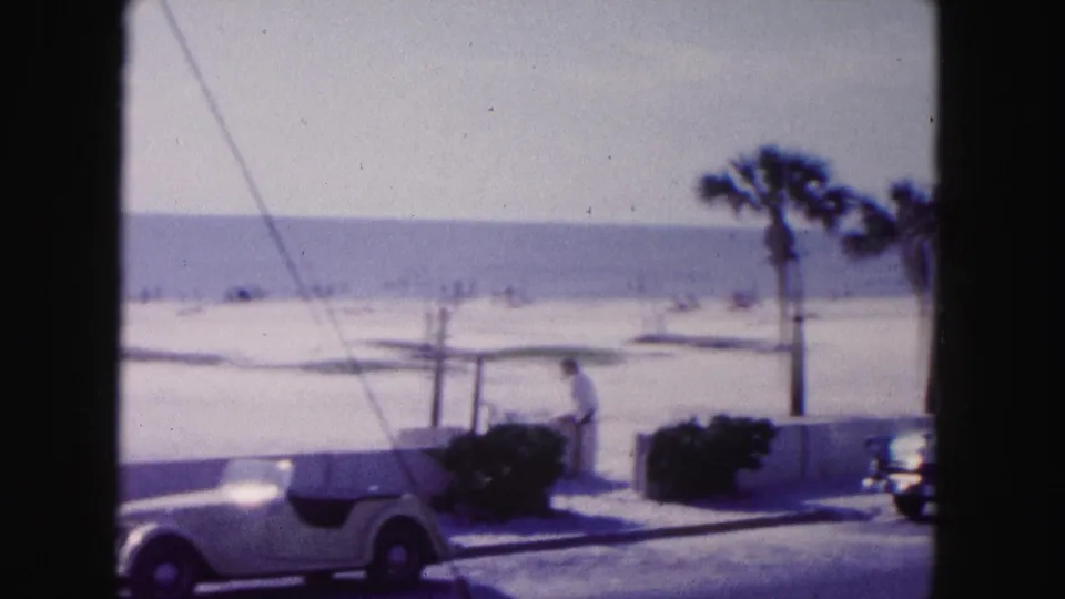 1960 Miami home-movie frame showing a beachfront road, white convertible, palms, low seawall, beach, ocean, and a distant person walking.