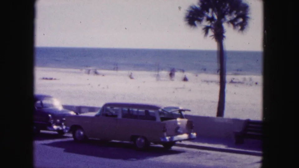 1960 Miami beach view with mid-century cars parked by a low wall, a palm tree, sandy beach, ocean, and small figures in the distance.