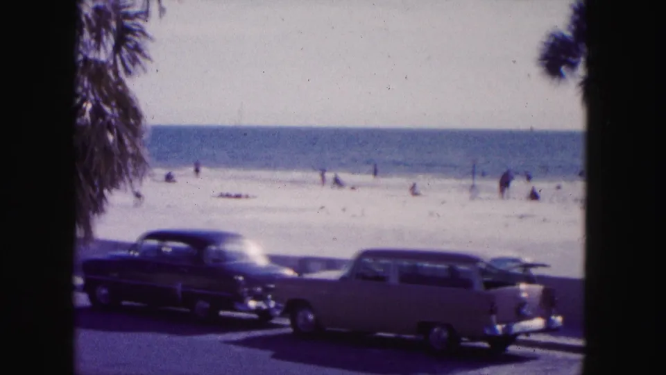 1960 Miami home-movie frame of a dark sedan and light station wagon on a seaside road, with palms, beach, ocean, and distant people behind them.