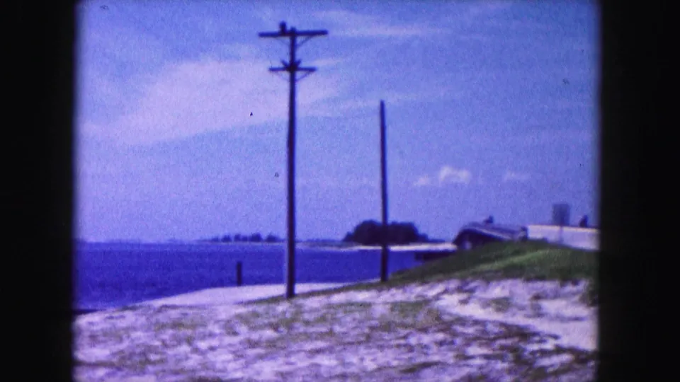 Cutaway to a Miami waterfront view with utility poles, shoreline, water, and distant low buildings in 1960 8mm footage.