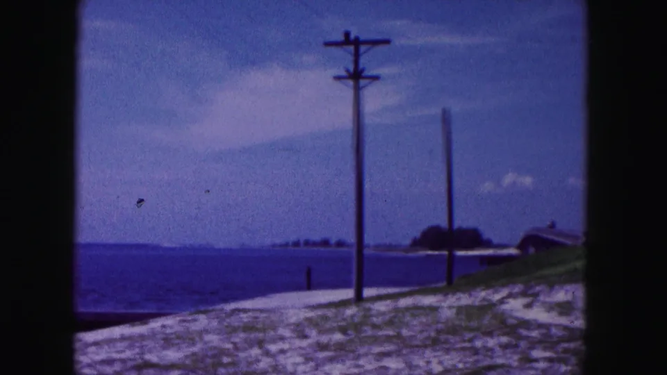 1960 Miami-area 8mm frame showing a utility pole on a grassy sand edge beside ocean water and a distant shoreline form.