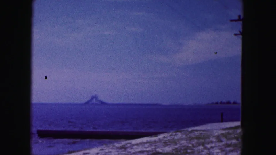 1960 Miami-area 8mm view looking over sand and ocean toward a distant landform or structure, with utility pole edges near the frame.