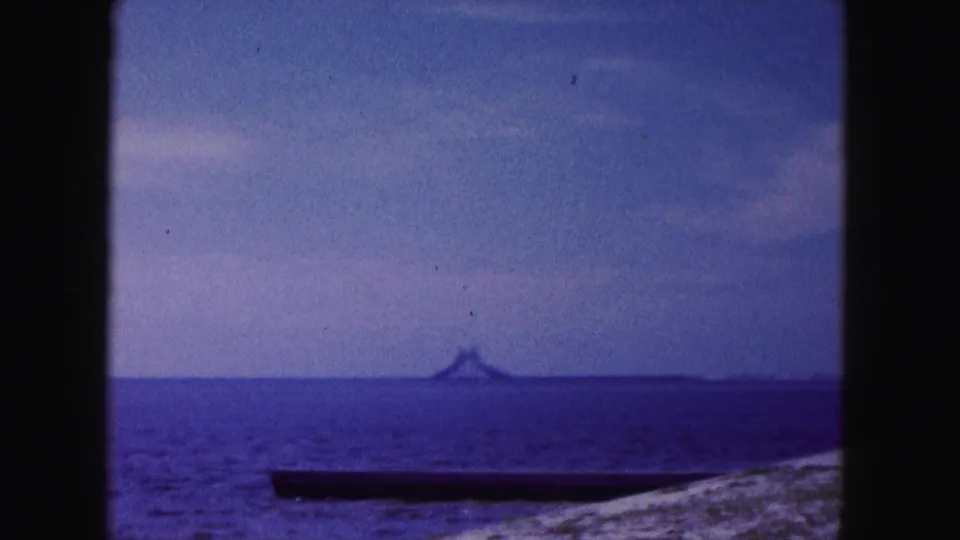 1960 Miami-area coastal frame with blue ocean, pale sand, a narrow dark shoreline object, and a distant landform or structure.