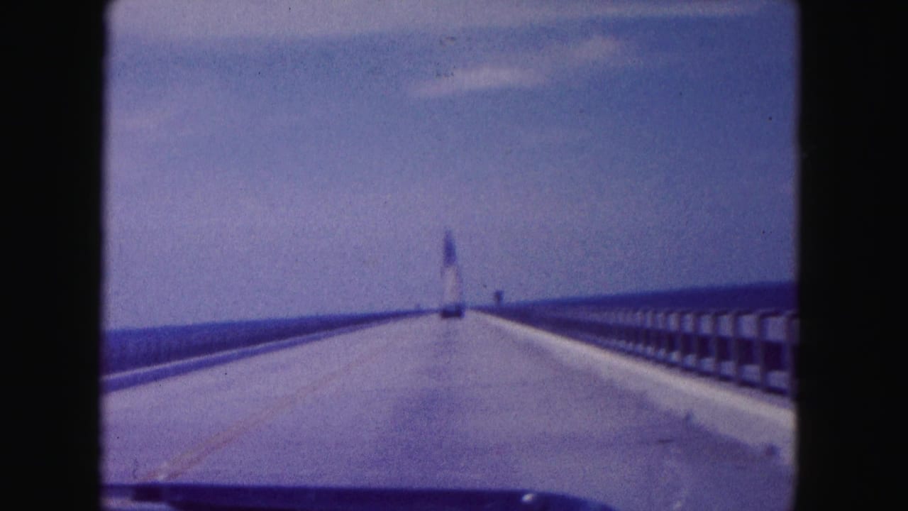 1960 Miami home-movie view from a car crossing a long bridge toward a distant tower-like structure under blue sky.