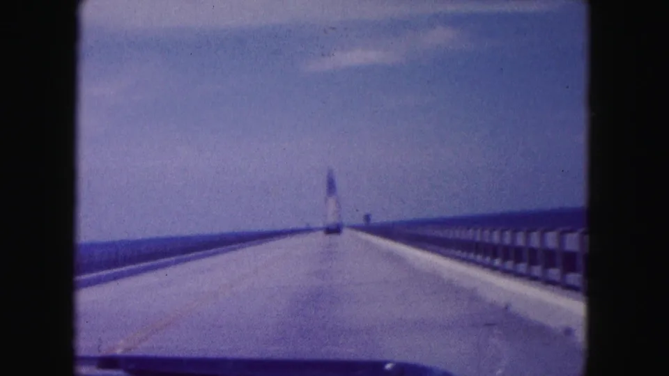 1960 Miami home-movie view from a car crossing a long bridge toward a distant tower-like structure under blue sky.