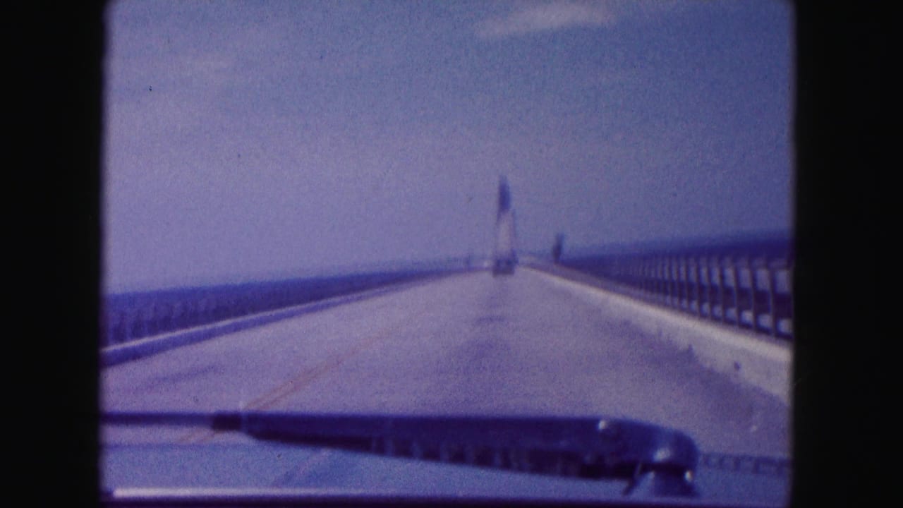 Car windshield view on a Miami bridge in 1960, with guardrails, open sky, distant tower, and wiper visible in front.