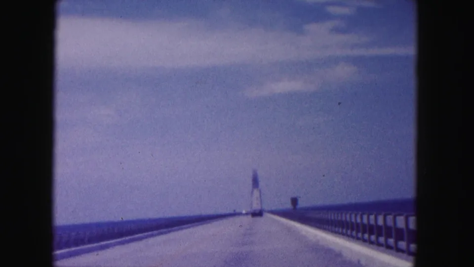 Final 1960 Miami bridge view from a moving car, showing guardrails, open water, clear sky, and the distant tower structure.