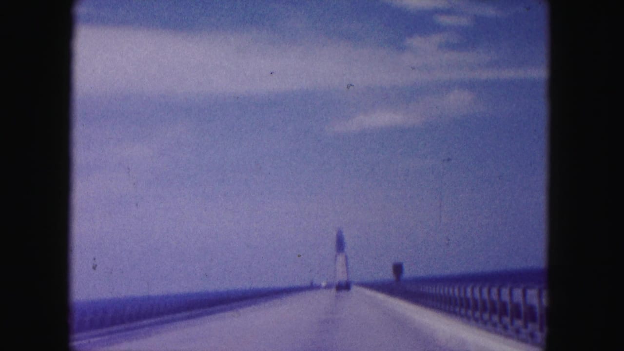 Roadway on a long bridge seen from a moving car, with railings and a distant tower shape ahead.