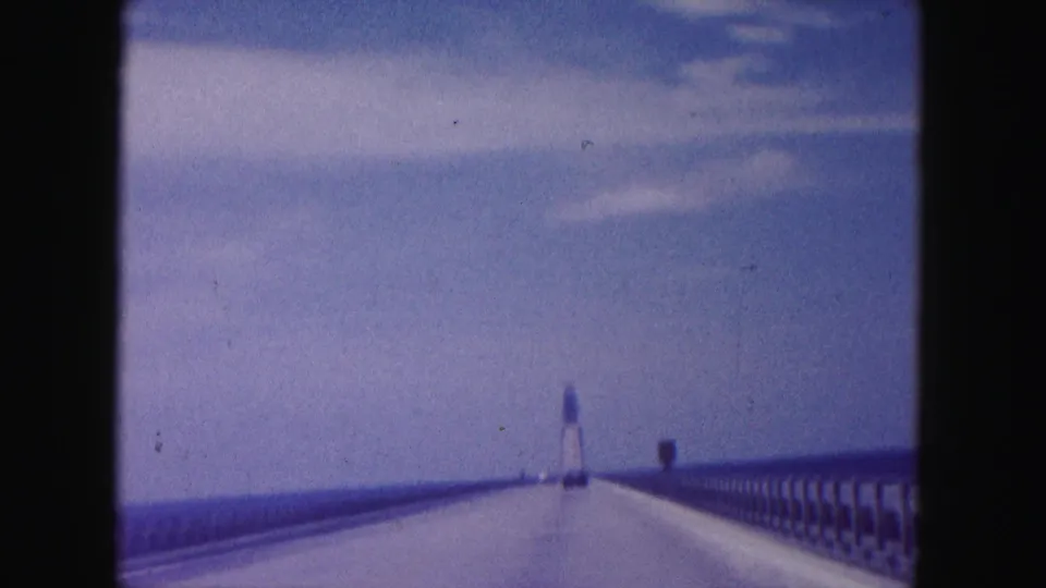 1960 Miami-area 8mm home-movie frame showing a car viewpoint on a long bridge over water, approaching a distant tower-like structure.