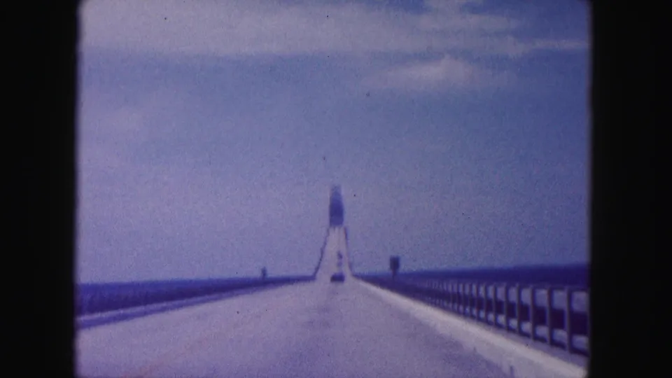 Forward-facing 8mm frame of a 1960 Miami-area bridge crossing, with open water beside the road and a tower-like form ahead.
