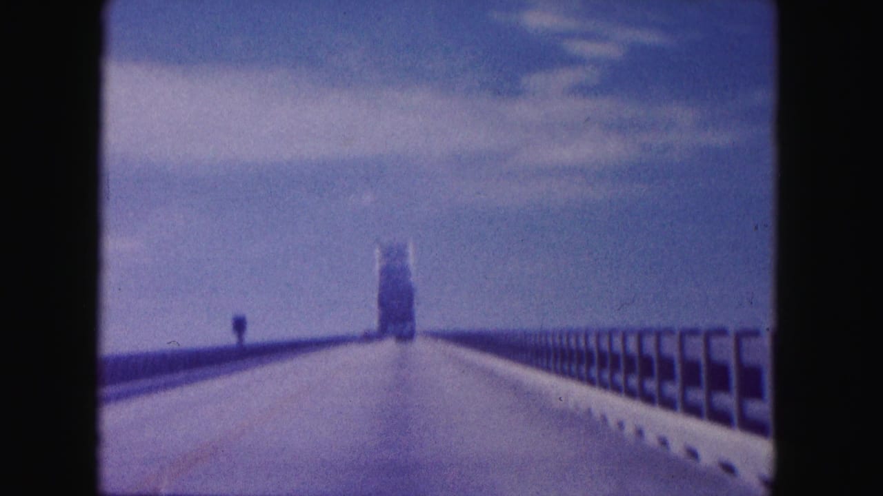 Passenger-view 8mm frame of a Miami bridge drive, with railings on both sides and a tower ahead in soft focus.