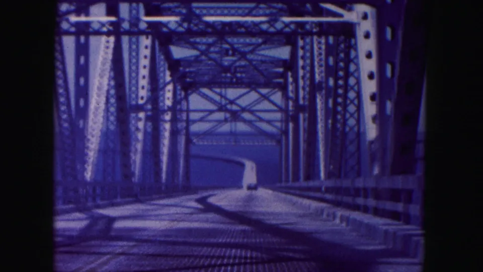 1960 Miami-area 8mm frame showing a vehicle-point-of-view drive through a steel truss bridge with water beyond.