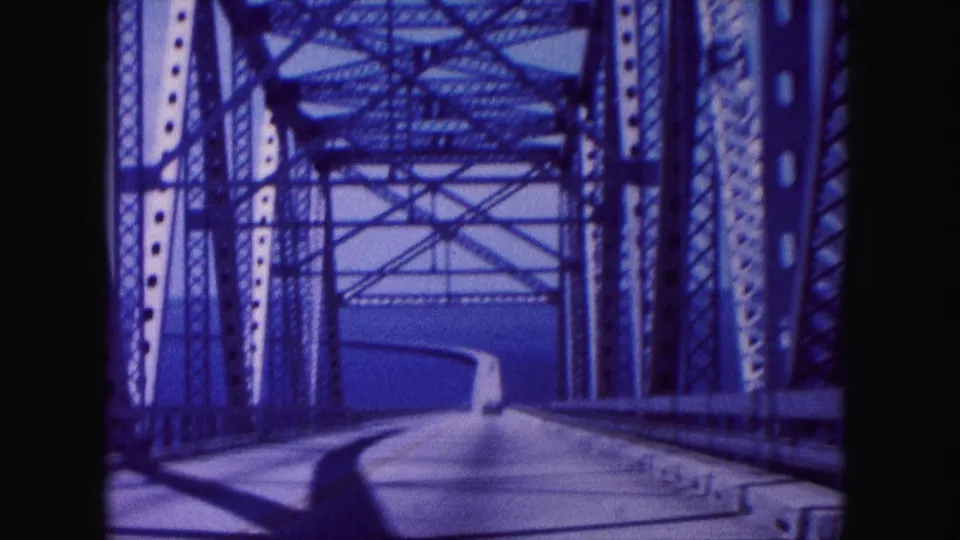 Vehicle POV on a Miami, Florida bridge in 1960, with truss beams framing a bright roadway over water.