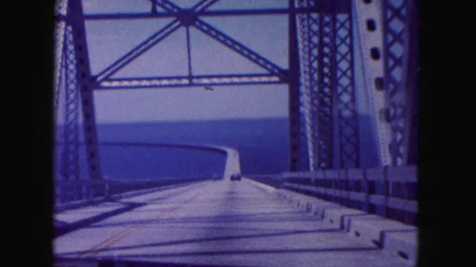 Final 1960 Miami bridge-driving frame showing steel supports, a curving road deck, and broad water beyond.
