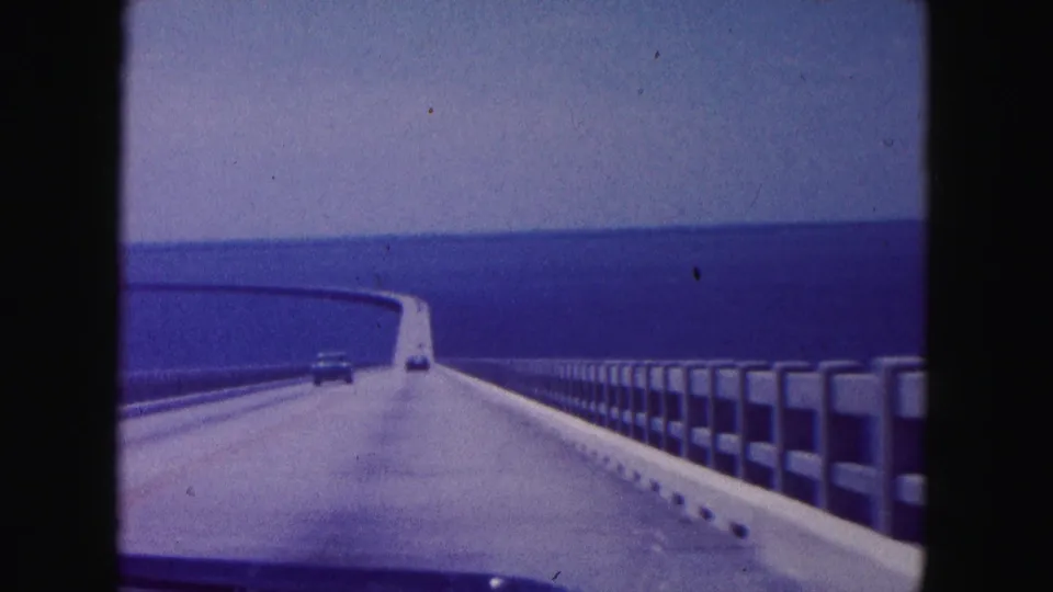 1960 home-movie view from a moving car crossing a long Miami-area bridge over open water, with railings and a distant vehicle ahead.