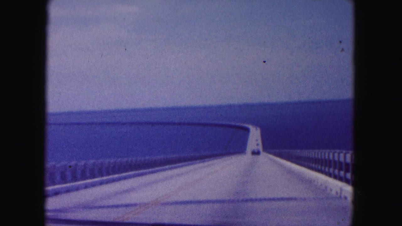 Forward-facing 8mm frame of a car crossing a curving bridge over water in the Miami, Florida area, with a vehicle visible ahead.