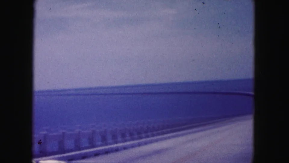 Soft 8mm frame from a moving car on a long bridge, showing railing, roadway, open water, and pale sky in a Miami-area travel view.