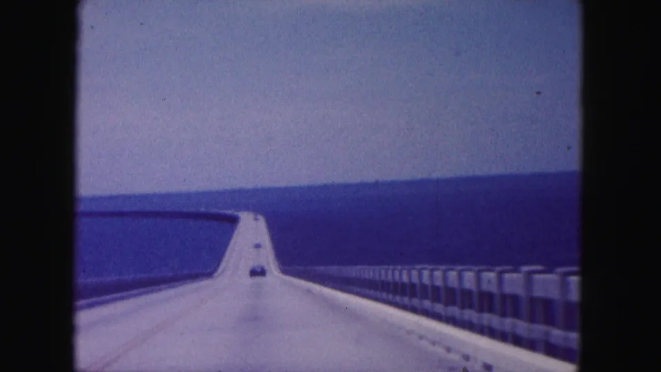 1960 8mm travel frame looking down a long bridge over water near Miami, with concrete barriers, railings, and a distant car ahead.