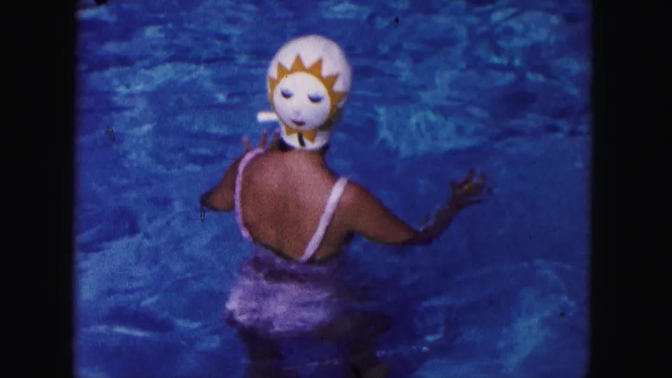 1960 Miami home-movie frame of a swimmer in blue pool water wearing a sun-shaped mask, white cap, and vintage swimsuit.