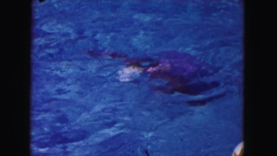 1960 Miami home-movie frame of a swimmer partly underwater in a blue pool, with the white cap visible near the surface.