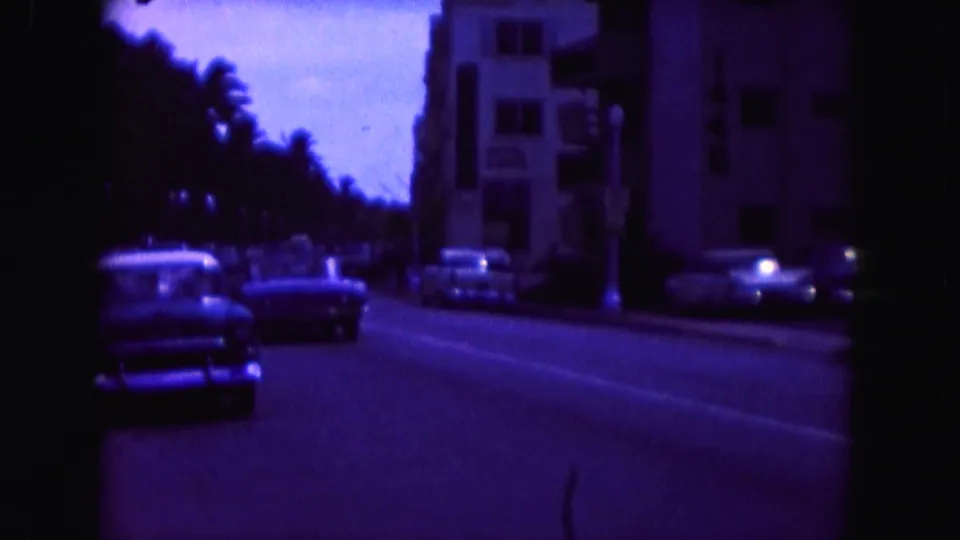 1960 Miami street view with classic cars moving along an avenue beside parked vehicles and mid-century buildings.