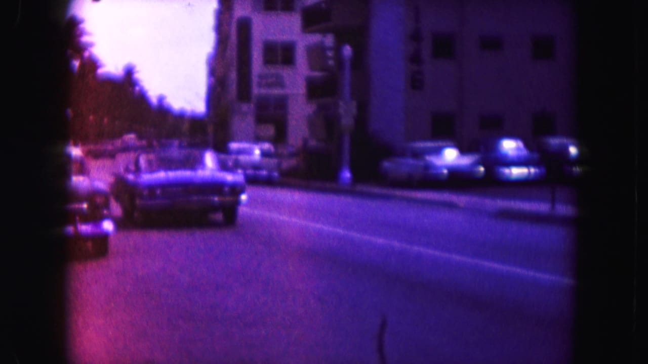 Car approaching on a purple-magenta street with parked cars and apartment buildings nearby.