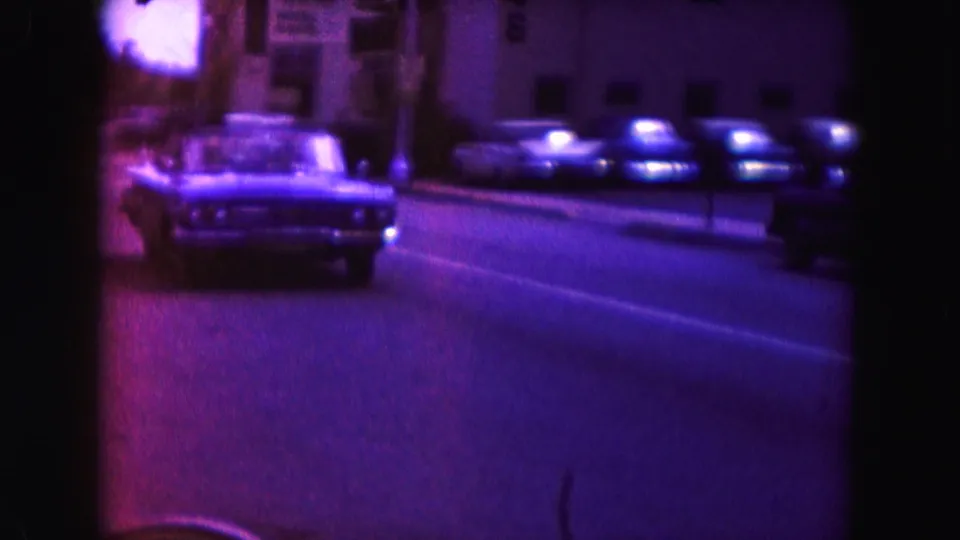 A classic American car passes closer to the 8mm camera on a Miami street lined with parked vehicles and buildings.