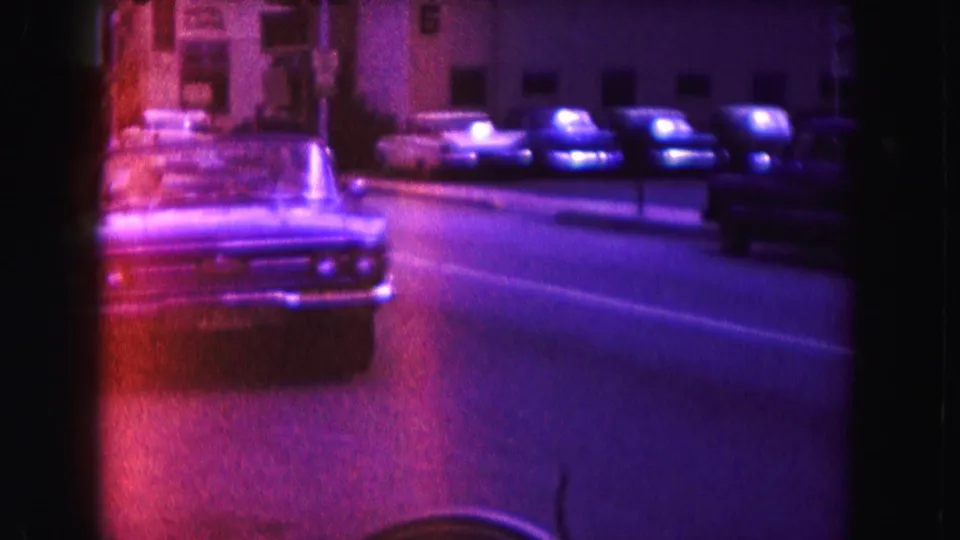 Rear view of a classic car moving along a 1960 Miami avenue beside parked cars and mid-century apartment buildings.
