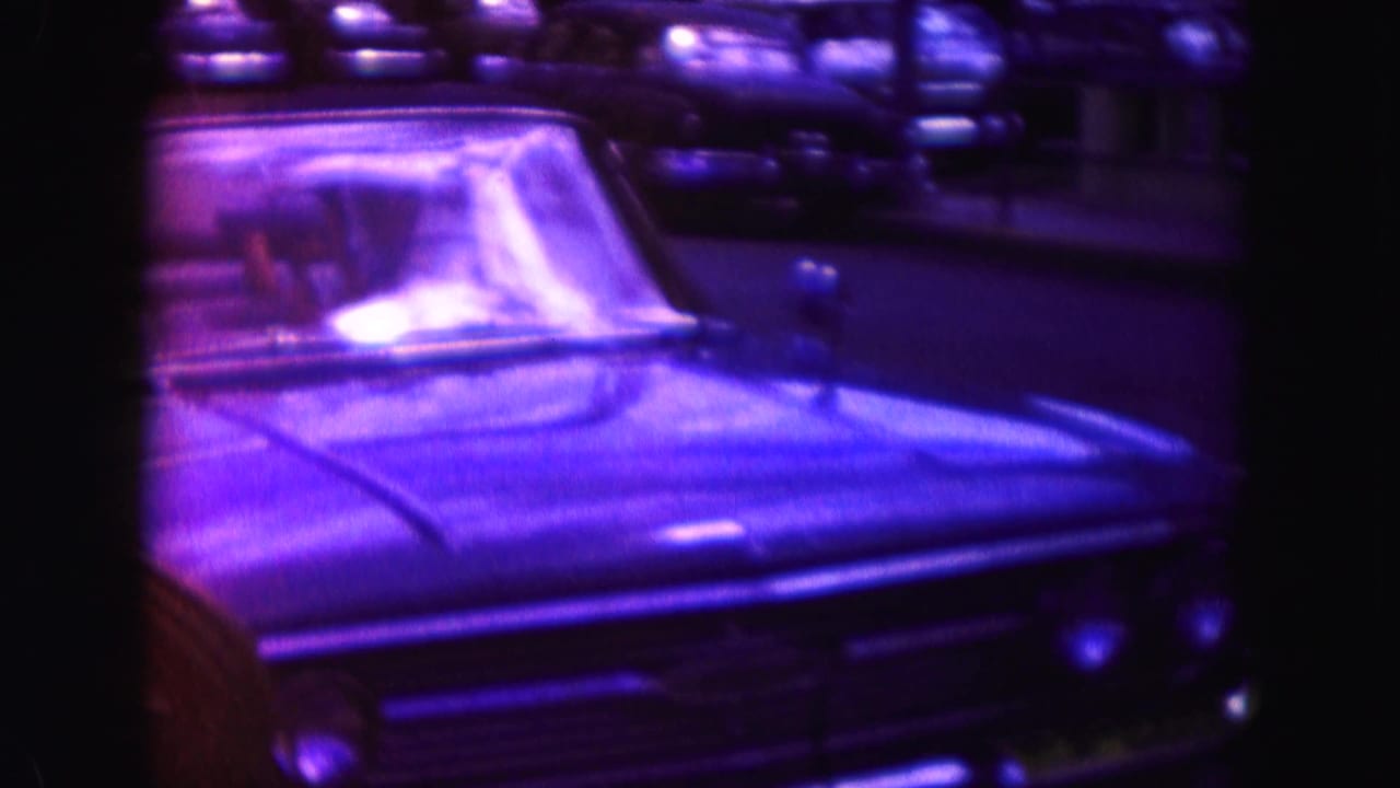 Classic convertible seen close from the front at night, with windshield glare and parked cars behind it in Miami home-movie footage.
