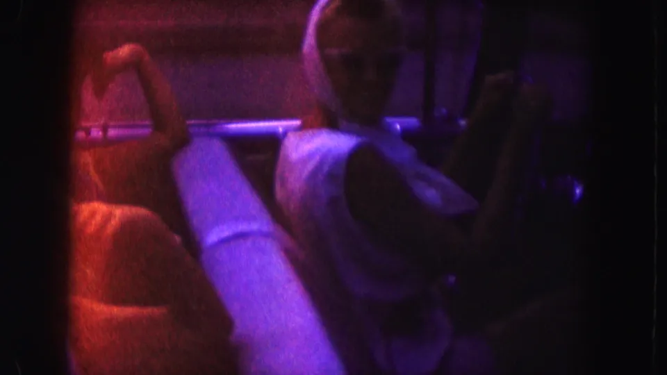 Passengers sit inside an open convertible in the 1960 Miami street scene, with faces softened by low light and color-shifted 8mm exposure.