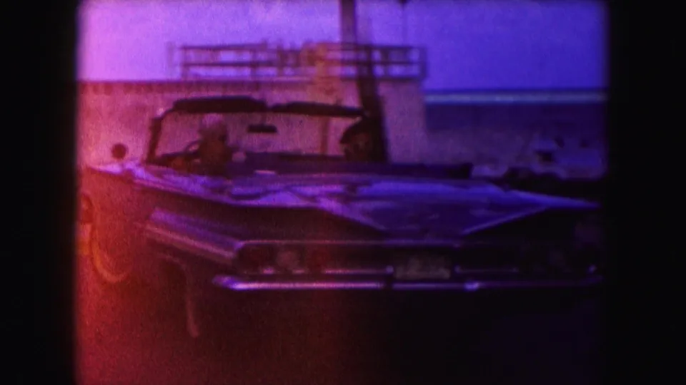 A 1960s convertible is seen from the front as it moves along a Miami coastal road, with passengers visible through the windshield.