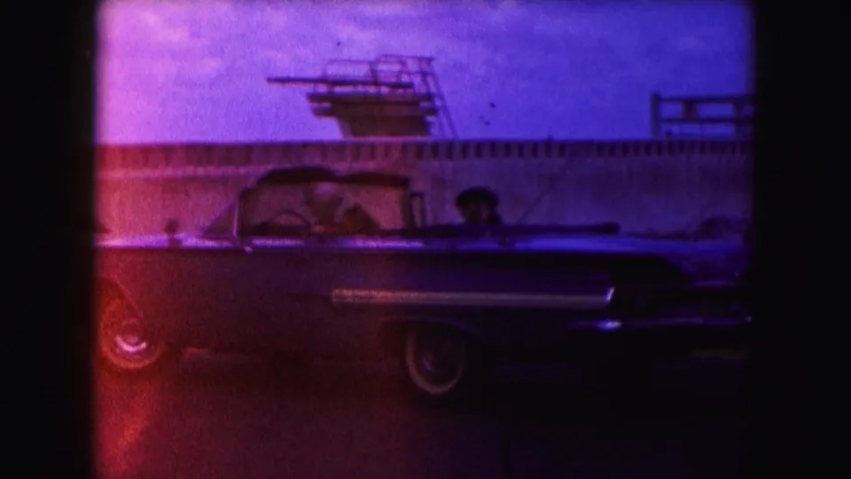 Side profile of a classic convertible in Miami, with passengers visible and a waterfront railing or pier structure behind it.