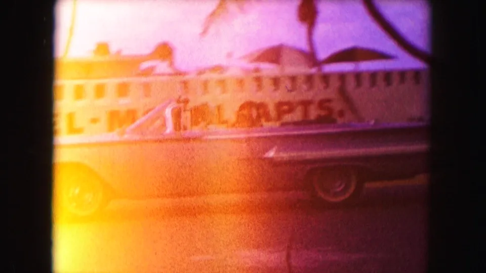 1960 Miami frame with a classic convertible moving past hotel-motel signage under heavy orange exposure bloom.