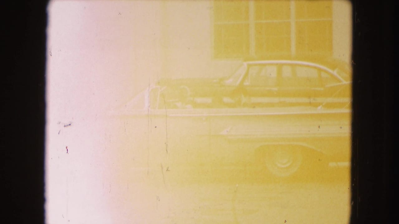 Faint view of a large blue convertible in metadata-supported 1960 Miami, seen beside a building through yellowed, overexposed 8mm film.