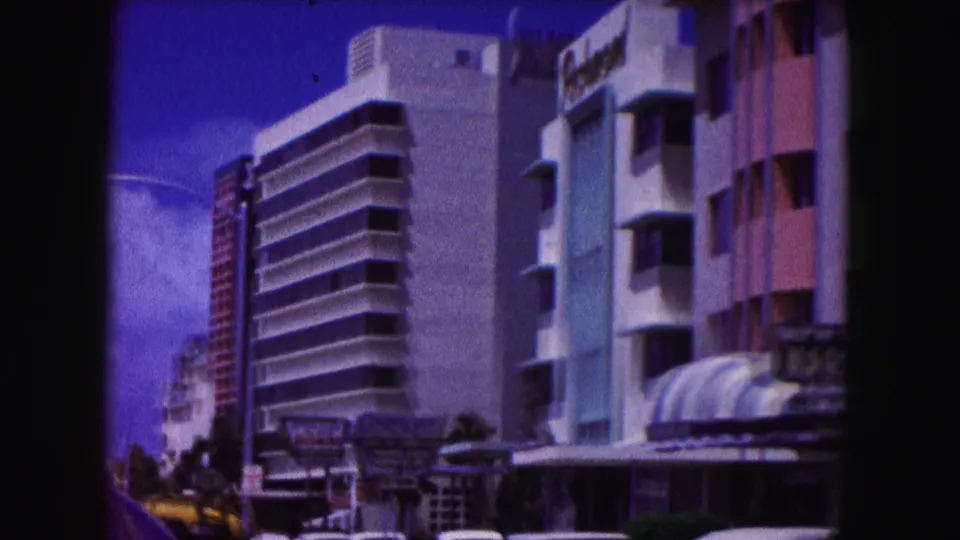1960 Miami 8mm view looking down a hotel-lined street with a white modern facade, pastel building fronts, signage, awnings, and cars.