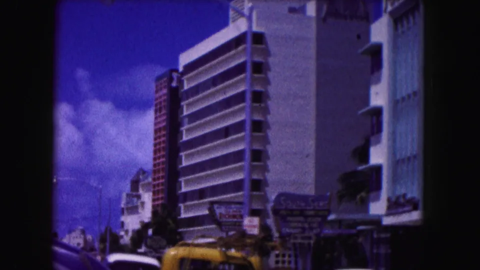 1960 Miami street frame with a tall balcony-front hotel, visible roadside signs, yellow and white cars, palm greenery, and bright blue sky.